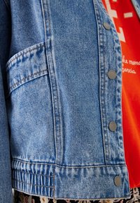 Džínová bunda světle modré barvy s kovovými stříbrnými cvoky, prošitými detaily a boční kapsou. Pod bundou jasně oranžové tričko.