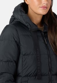 Giacca a vento nera con chiusura a zip, cappuccio e tessuto testurizzato. Presenta una fascia con logo sul davanti e un design trapuntato cucito.