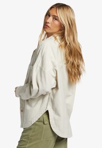 Light beige button-up shirt featuring a relaxed fit, curved hem, and long sleeves, paired with green cargo pants. Soft texture and minimal design.