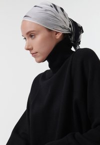 Pull en maille noire à col montant, avec une coupe ample, accompagné d'une écharpe grise clair ornée de rayures abstractes noires, nouée dans le dos.
