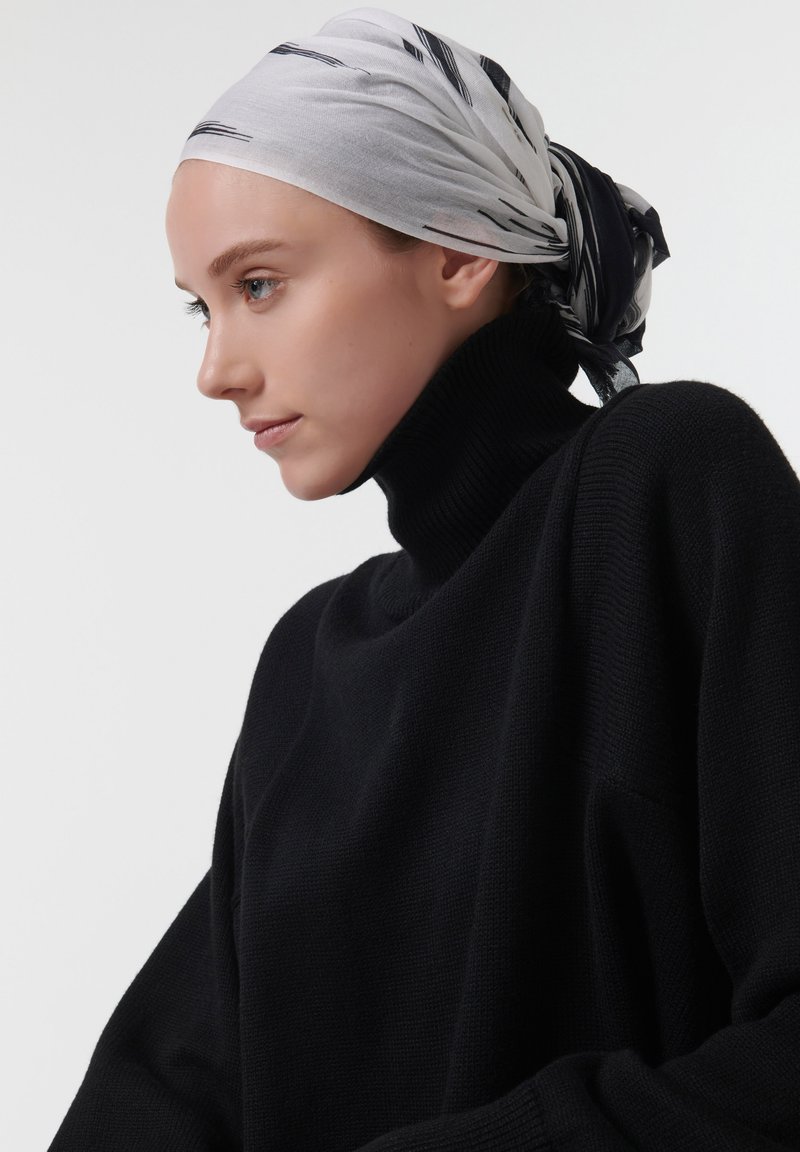 Pull en maille noire à col montant, avec une coupe ample, accompagné d'une écharpe grise clair ornée de rayures abstractes noires, nouée dans le dos.