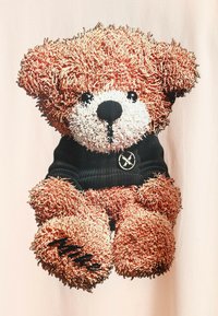 Diseño de oso de peluche marrón con pelaje texturizado, que lleva puesto un suéter negro de canalé con un logotipo. Impreso sobre un fondo de tela de color claro.