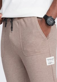 Beżowe, tkane spodnie z elastycznym paskiem w talii i sznurkiem. Posiadają boczną kieszeń naszywaną z metką marki; gładka tekstura. Widoczny czarny zegarek.