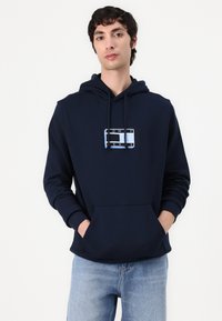 Felpa blu navy con tasca frontale, caratterizzata da una grafica rettangolare in blu chiaro e bianco. Il materiale sembra morbido e dal texture piacevole.