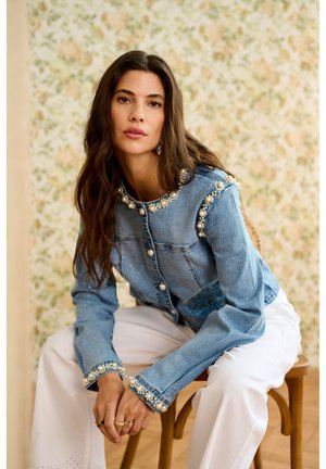 Pikkade juustega naine istub puidust toolil, kandes sinist pärlitega kaunistatud teksatagi ja valgeid pükse, taga lilleline tapeet.