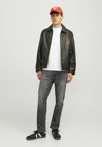 Veste en cuir noir, t-shirt blanc, jeans gris et baskets noires avec des rayures blanches. Casquette rouge avec détails textuels. Mains dans les poches.