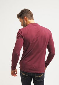 Camisa de manga larga color marrón oscuro con escote redondo, textura suave y diseño ajustado, combinada con jeans de mezclilla oscura con bolsillos traseros.
