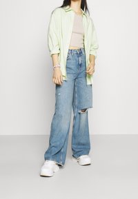 Jasnozielona koszula z długim rękawem na beżowym topie, zestawiona z przetartymi, luźnymi niebieskimi dżinsami i białymi trampkami. Różowa bransoletka na nadgarstku.