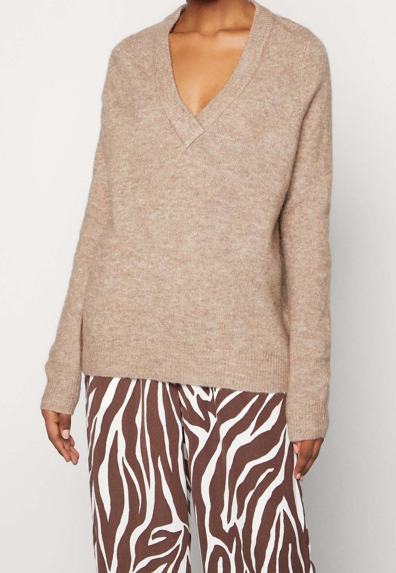 Beige V-Ausschnitt-Pullover aus weichem Stoff mit langen Ärmeln. Kombiniert mit braun-weißen, zebra-gemusterten Hosen im lässigen Schnitt.