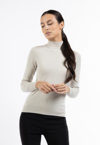 Pulover bej cu guler înalt dintr-un material moale, tricotat fin, cu mâneci lungi și un design ajustat, asortat cu pantaloni negri.