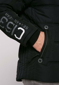 Giubbotto trapuntato nero con tasca con zip e polsini elasticizzati. La manica sinistra presenta un grande marchio bianco. Tessuto testurizzato con finitura opaca.