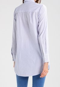 Camisa a rayas de botón en azul claro y blanco, con mangas largas, cuello de solapa y un corte holgado. Textura de tela suave con un dobladillo curvado.