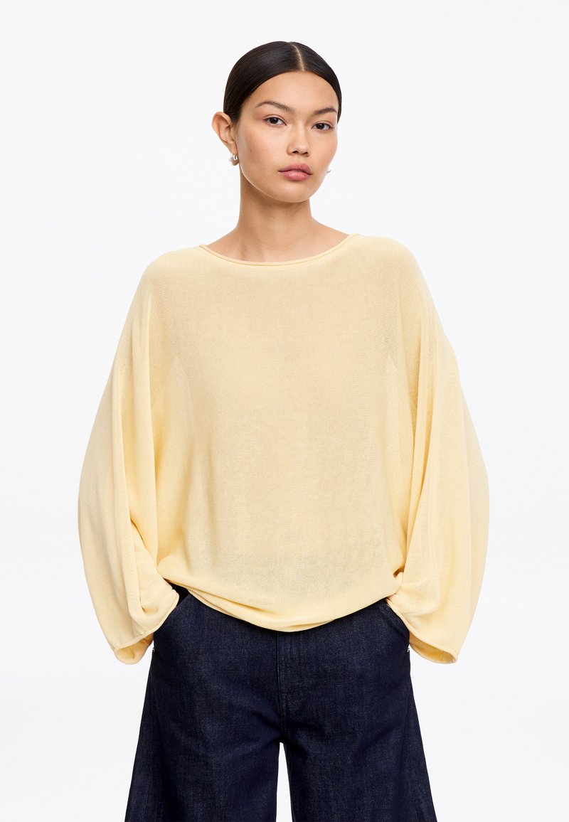 Femme aux cheveux foncés en chignon bas, portant un haut ample à manches longues jaune clair et un pantalon large sombre, mains dans les poches, fond neutre.