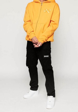 Sudadera amarilla con cremallera y bolsillo delantero con detalle gráfico, combinada con pantalones cargo negros con parche de logo lateral y zapatillas blancas.