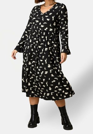 Vrouw draagt een zwarte midi-jurk met witte abstracte print, lange mouwen en zwarte robuuste enkellaarzen, staand tegen een lichte achtergrond.