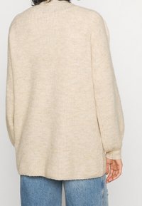 Beige pletený svetr s volným střihem, ribovanou strukturou, dlouhými rukávy a vysokým výstřihem, doplněný světle modrými džíny.