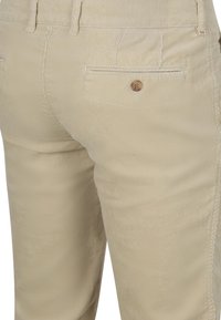 Beige corduroy broek met een achterzak met knoop en taillebandlussen, gezien vanaf de achterkant.