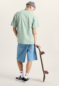 Mintgrøn bomulds T-shirt i løs pasform, parret med lyse denimshorts. Sort skateboard med røde hjul, slidte sorte sneakers med hvide strømper.