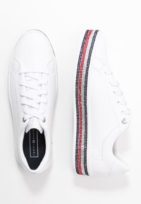 Białe skórzane sneakersy z płaskim zapięciem na sznurowadła, srebrnymi oczkami i dekoracyjnym teksturowanym paskiem w kolorach czerwonym, białym i niebieskim wzdłuż podeszwy.