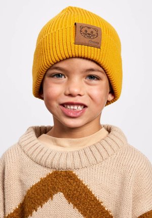 Smiling child wearing a yellow knit beanie with a tiger patch and a beige sweater with a brown chevron pattern.