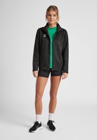 Zwarte zip-up jas met hoge kraag, gecombineerd met een groene shirt en zwarte shorts. Model draagt zwarte sportschoenen met witte sokken.