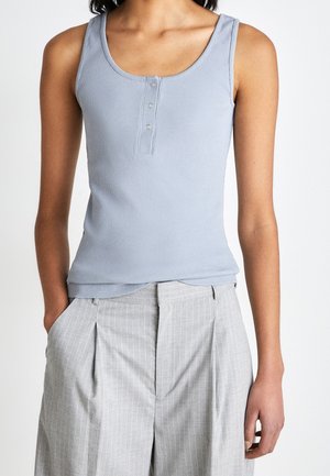 Femme portant un débardeur côtelé sans manches bleu clair avec des boutons, associé à un pantalon taille haute gris clair à fines rayures, mains dans les poches.
