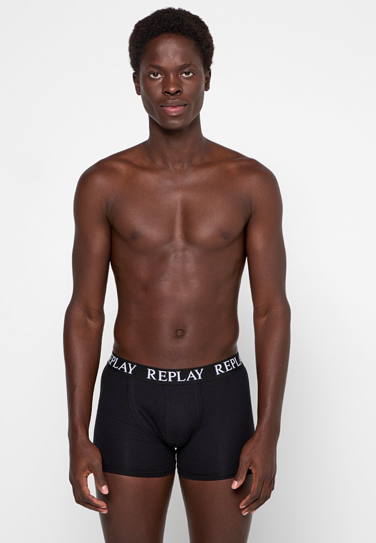 Junger Mann mit dunkler Haut, der schwarze Boxershorts mit "REPLAY" am Bund trägt, vor einem schlichten weißen Hintergrund stehend.