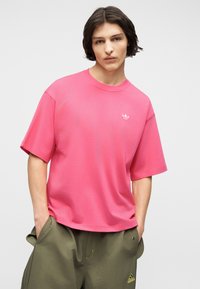 Νεαρό άτομο με σκουρόχρωμα μαλλιά μέχρι τους ώμους, φοράει φωτεινό ροζ κοντομάνικο μπλουζάκι Adidas και χακί παντελόνι Adidas, με τα χέρια στις τσέπες.