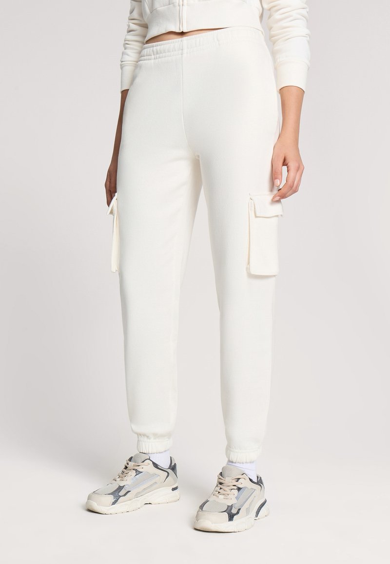 Białe dresowe spodnie wykonane z miękkiego materiału, z bocznymi kieszeniami cargo i elastycznymi mankietami, w zestawie z wielokolorowymi trampkami sportowymi.