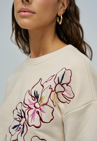 Mujer que lleva un sudadera beige con grandes flores bordadas en rosa y amarillo en el hombro, y pendientes de aro dorados.
