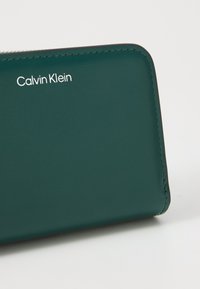 Green leather wallet with silver "Calvin Klein" logo, featuring smooth texture and precise stitching along the edges.