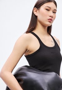 Μαύρο ριμπ τοπ με λεπτές τιράντες. Το μοντέλο κρατάει μια λεία, μαύρη τσάντα από δερματίνη. Μινιμαλιστικός σχεδιασμός χωρίς ορατά λογότυπα ή διακοσμήσεις.