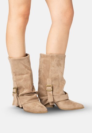 Ben iført midt på læggen brune ruskindsslouch-støvler med spidse tupper og dekorative gyldne spænder, på hvid baggrund.