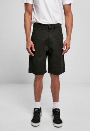 Mann trägt schwarze knielange Shorts, weiße Kniestrümpfe, schwarz-weiße Sneakers und ein weißes T-Shirt, sichtbar von der Taille bis zu den Knien.