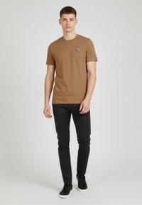 Camiseta de algodón marrón con cuello redondo y mangas cortas, que presenta un pequeño parche de logo negro en el pecho. Combinada con pantalones negros y deportivas.