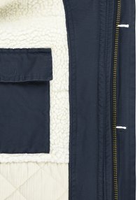 Veste matelassée bleu marine avec doublure en peluche beige, dotée d'une poche carrée et d'une fermeture éclair dorée. La texture contraste entre des matériaux lisses et doux.