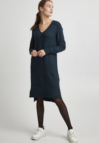 Robe en tricot bleu foncé avec un col en V, des manches longues, des fentes latérales et un ourlet légèrement plus long à l'arrière. Portée avec des collants noirs et des baskets blanches.