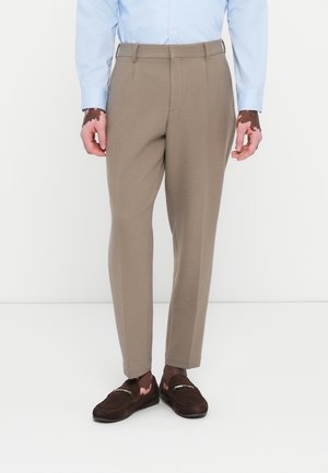 Beige slim-fit trousers with a smooth texture, flat front, and crease down the legs, paired with dark brown loafers and a light blue shirt.