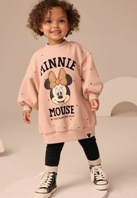 Ung barn med lockigt hår som bär en stor rosa Minnie Mouse-tröja, svarta leggings, beige strumpor och svarta högklackade sneakers.