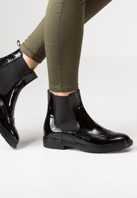 Botas Chelsea de charol negro con paneles elásticos laterales, combinadas con pantalones ajustados de color verde oliva, sobre un fondo claro y sencillo.