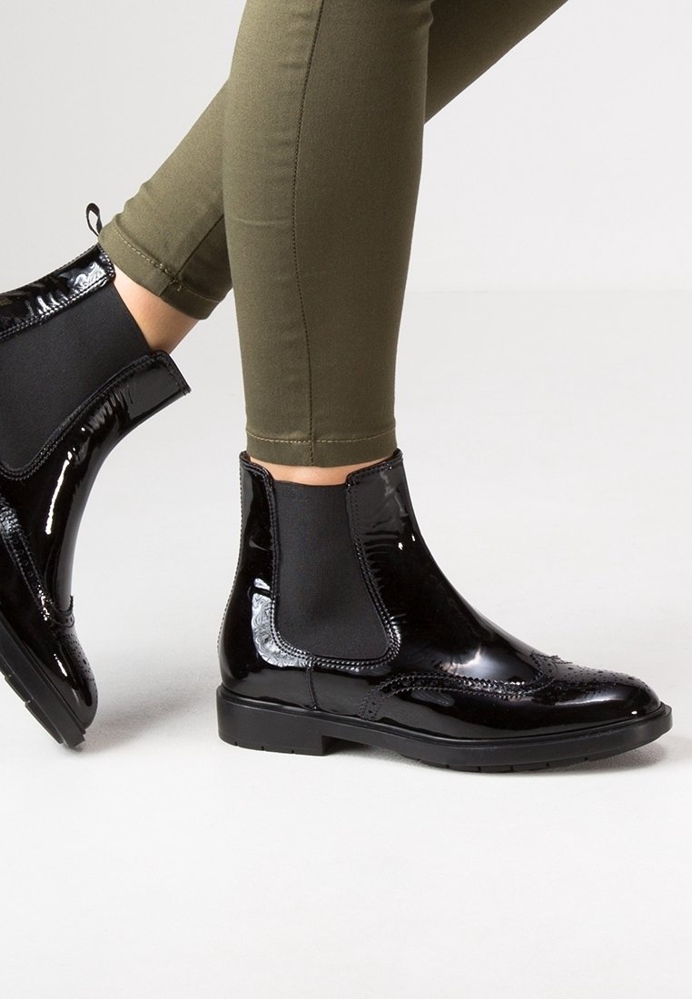Botas Chelsea de charol negro con paneles elásticos laterales, combinadas con pantalones ajustados de color verde oliva, sobre un fondo claro y sencillo.