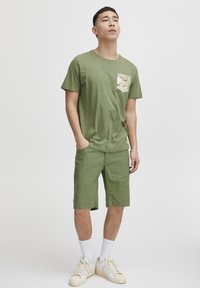 Alyvuogių žalios spalvos medvilninė marškinėliai su raštuotu kišene, derinami su atitinkančiais trumpais šortais. Dėvimi su baltais kulkšnies ilgio kojinėmis ir kreminiais sportbačiais.