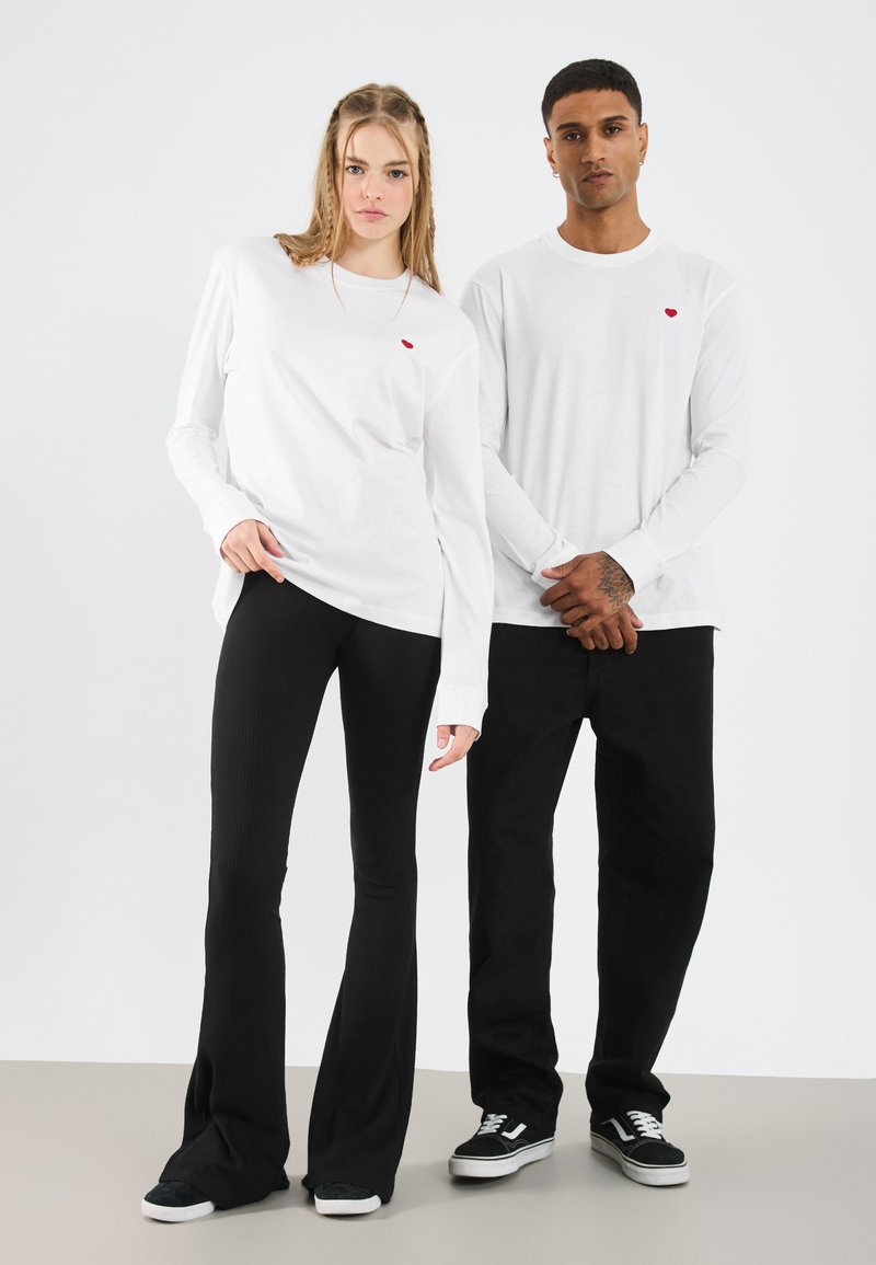 Λευκά πουκάμισα με μακριά μανίκια και λεπτομέρεια μικρής κόκκινης καρδιάς, συνδυασμένα με μαύρα κομψά παντελόνια με ραβδώσεις. Τα μοντέλα φορούν μαύρα αθλητικά παπούτσια, με φόντο ουδέτερης απόχρωσης.