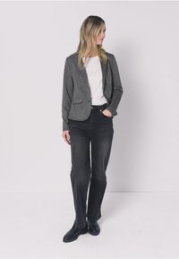 Donna in piedi con abbigliamento casual: blazer grigio, camicia bianca, jeans neri e scarpe nere, che guarda di lato contro uno sfondo semplice.