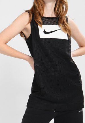 Mujer con top negro sin mangas de malla Nike con logo blanco, posando con las manos en las caderas sobre un fondo liso.