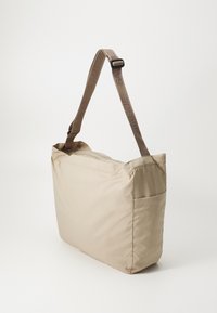 Beige stoff skulderveske med bred kropp, justerbar brun stropp og sidelomme. Glatt tekstur med enkle, rene linjer.