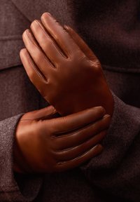 Braune Lederhandschuhe mit einer glatten Textur, die vier verlängerte Finger und ein schlichtes Design aufweisen. Sie werden vor einem dunklen Hintergrund zusammengehalten.