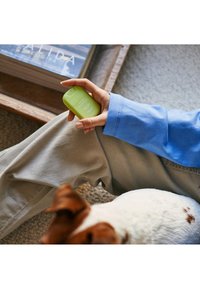 Spray verde tenuto in mano, con finitura opaca e testo bianco. Camicia a maniche lunghe blu chiaro e pantaloni beige visibili. Cane in primo piano.