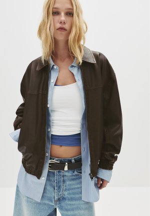 Bruine ritsjack met elastische mouwen over een blauw-wit gestreept shirt. Witte tanktop eronder, gecombineerd met blauwe en versleten denim jeans.