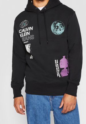 Personne portant un sweat à capuche noir Calvin Klein avec des graphismes et un texte sur le thème de l'espace, associé à un jean bleu.